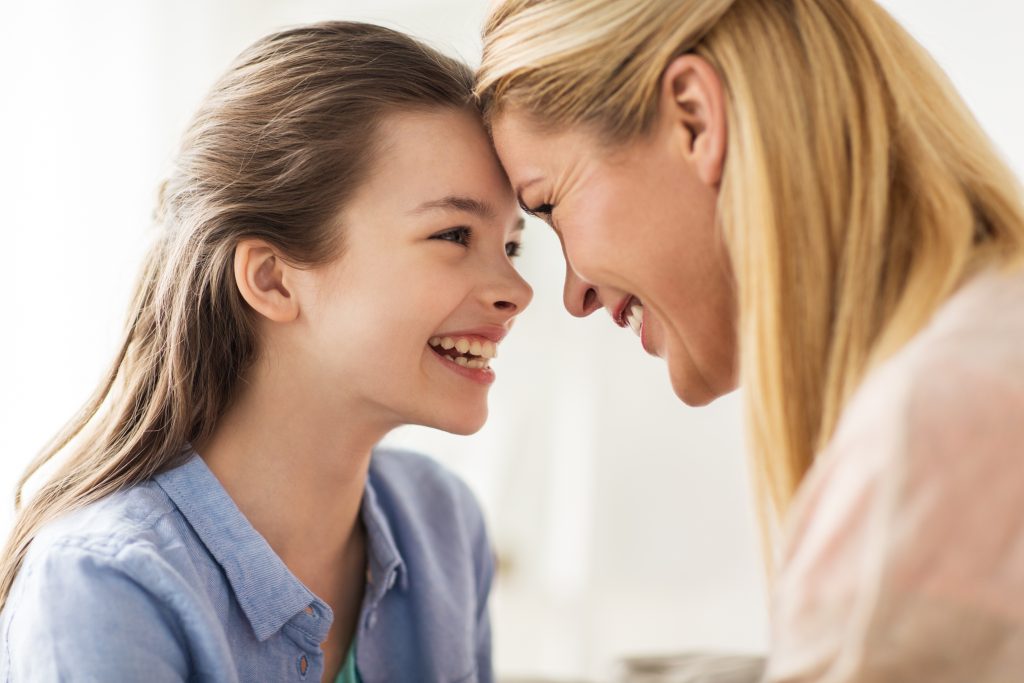Une mère et sa fille se regardant dans les yeux en riant, front contre front, partageant un moment de complicité et de tendresse. La petite fille est brune avec une chemise bleue et la maman est blonde et porte une chemise ose claire