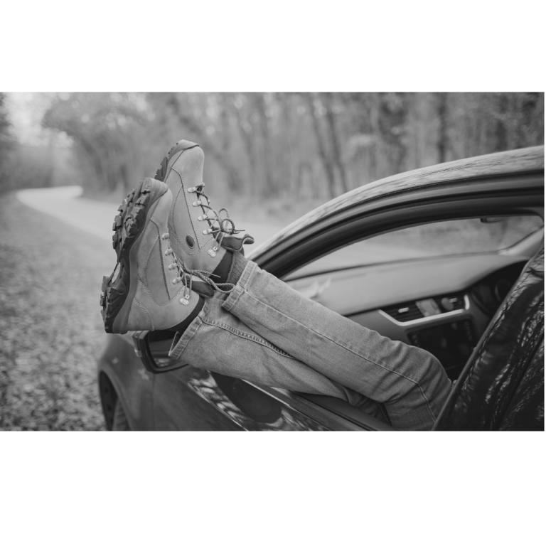 Pieds avec chaussures de randonnée sortant par la fenêtre d'une voiture, sur une route forestière en noir et blanc, moment de détente et d'évasion en pleine nature