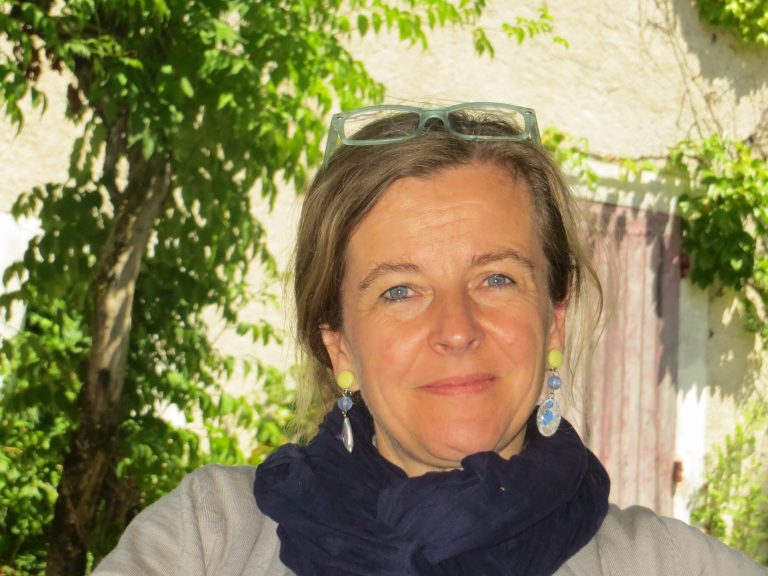 Portrait d’une femme souriante aux yeux bleus, portant une écharpe bleu marine, des boucles d’oreilles colorées et des lunettes posées sur la tête, en extérieur devant un mur clair et végétation