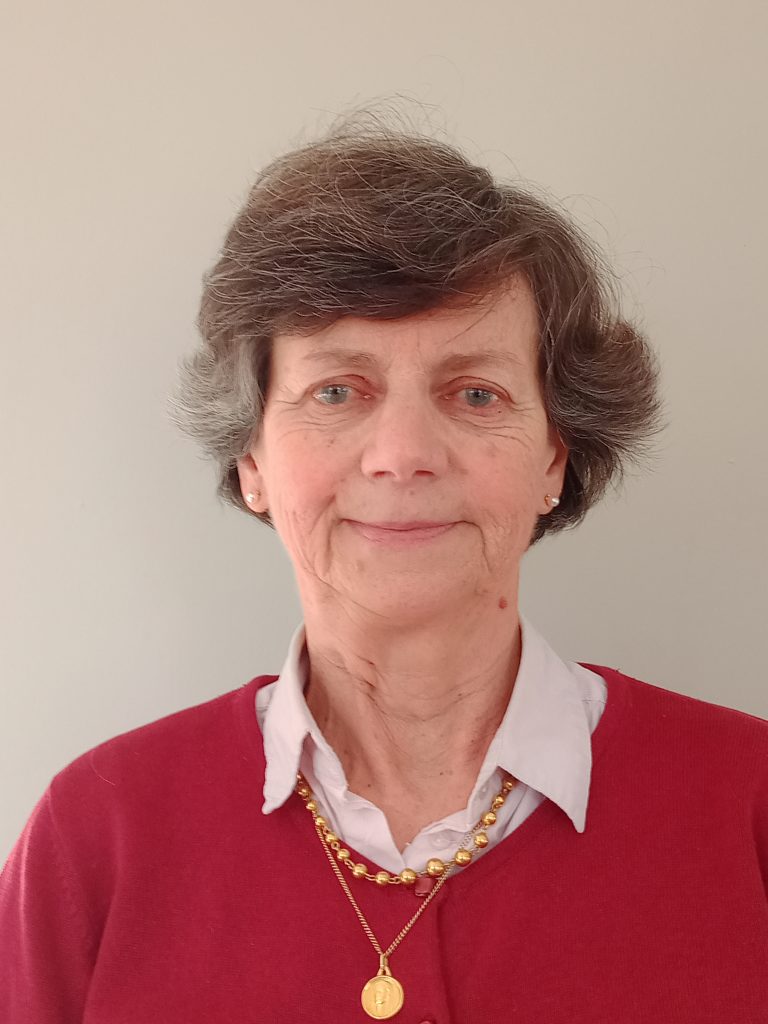 Anne a un léger sourire sur les lèvres/ Elle est blanche de peau avec des cheveux courts de couleurs brun et gris. Elle porte un gilet rouge part dessus une chemise blanche et des colliers en or.