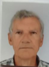 Portrait d'un homme aux cheveux gris courts, regardant directement la caméra avec une expression neutre, vêtu d'une chemise à motifs, sur un fond gris uni.