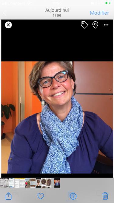 Portrait d'une femme souriante et chaleureuse avec des lunettes noires e une coupe de cheveux courte et brune. Elle porte un foulard bleu et un chemisier de couleur violette