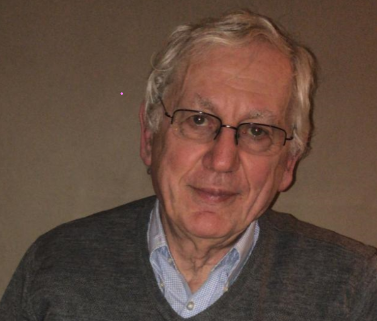 Portrait d’un homme senior aux cheveux blancs, portant des lunettes, une chemise bleue et un pull gris, posant devant un fond uni avec un léger sourire