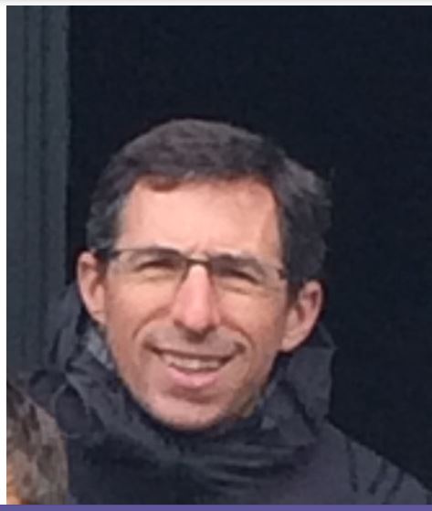 Portrait d'une homme avec un sourire, des lunettes noire rectangle et une coupe de cheveux courte de couleur foncée. Il porte également une veste imperméable.