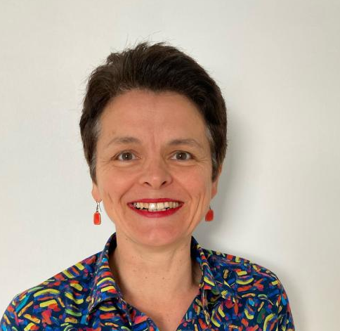 Portrait d’une femme souriante aux cheveux courts bruns, portant une chemise colorée à motifs et des boucles d’oreilles rouges, sur fond blanc
