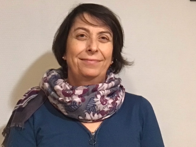 Portrait d'une femme joyeuse et souriante avec des cheveux noir et coupe courte arrivant au niveau des épaules. Elle porte également un foulard avec fleurs de couleur bleu, rose et violet. Elle port également un gilet bleu.